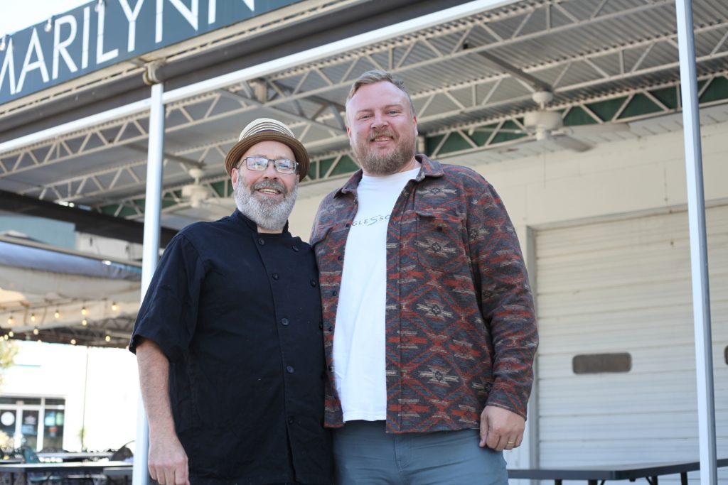 A horizontal photo shows two burly, light-skinned men posing for a casual portrait in front of a restaurant