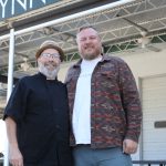 A horizontal photo shows two burly, light-skinned men posing for a casual portrait in front of a restaurant