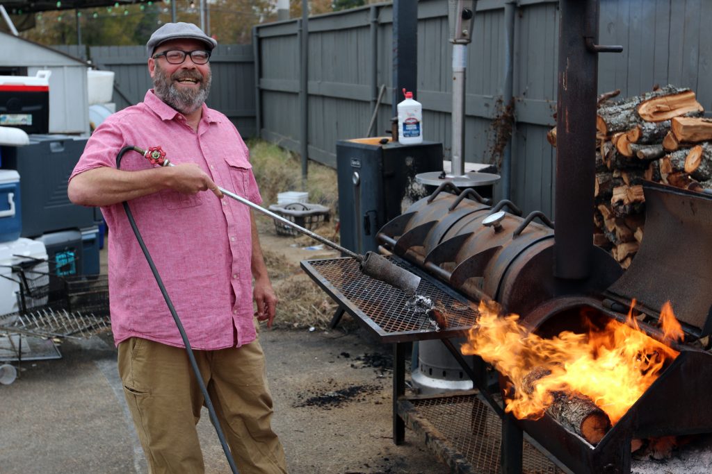 A horizontal photograph shows a man in a pink shirt smiling as he lights a grill with a flamethrower