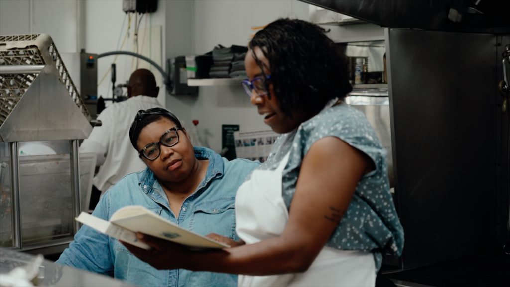 A photo shows two women standing in a kitchen, looking closely at a book