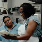 A photo shows two women standing in a kitchen, looking closely at a book