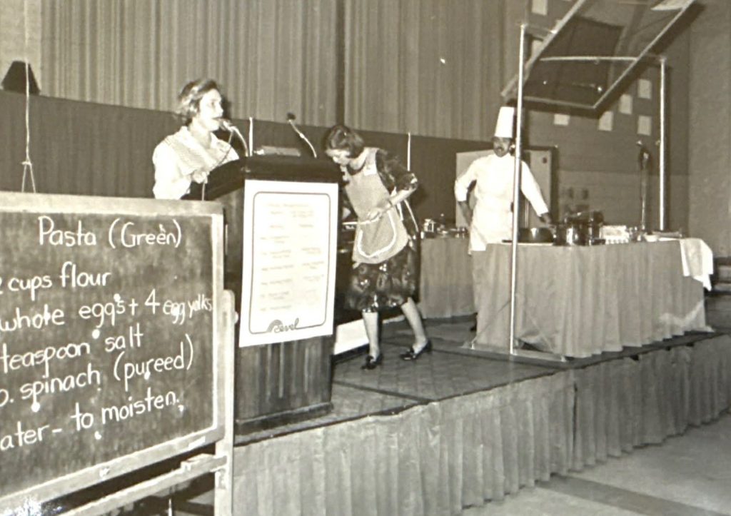 A black and white newspaper photo shows a woman cooking on a large stage